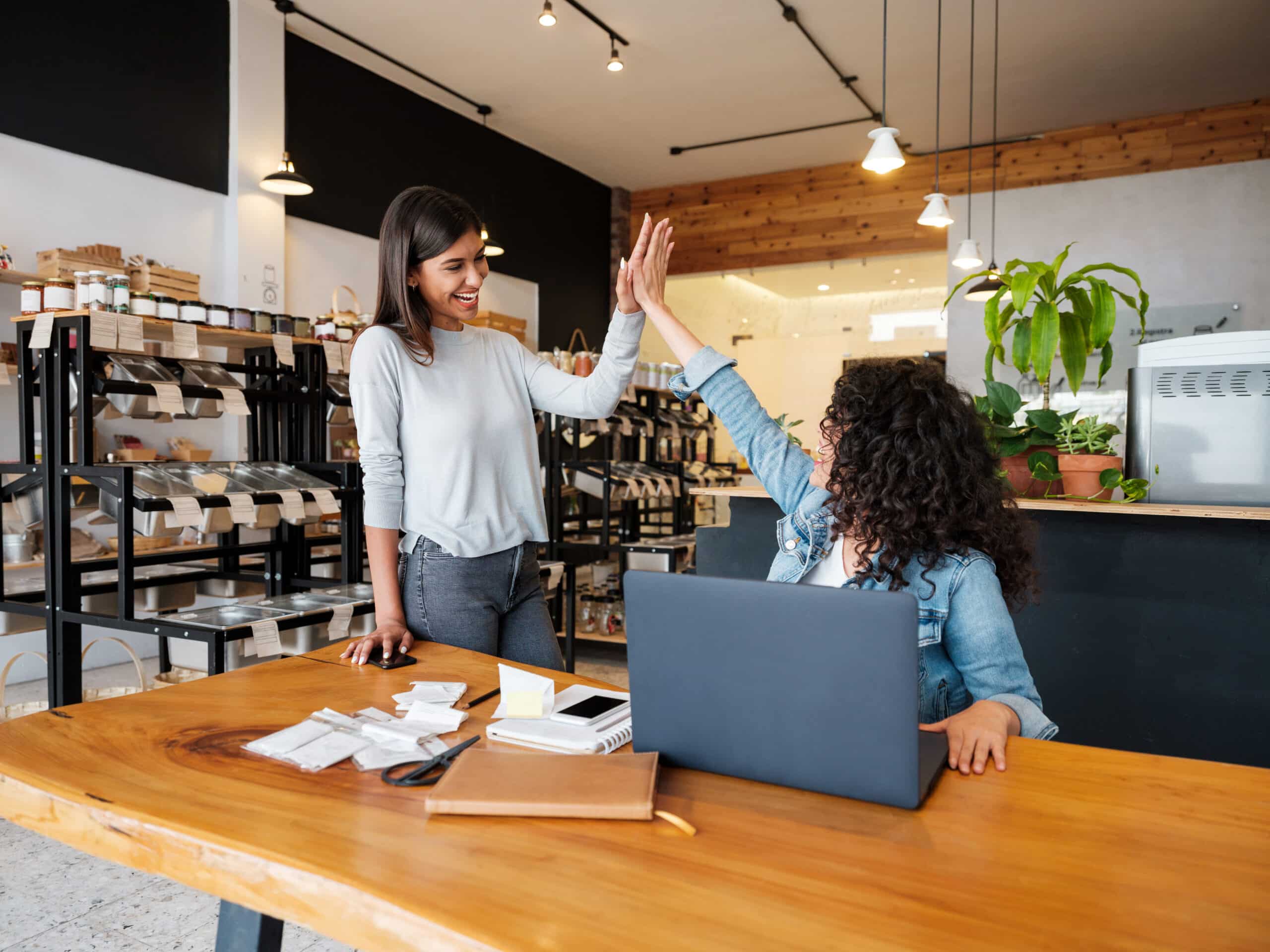 Two young Latina businesswomen in a small food shop giving high five and smiling at each other.
